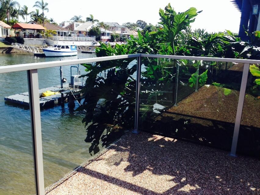 Waterfront patio with glass railing, potted plants, and a docked boat on a sunny canal.