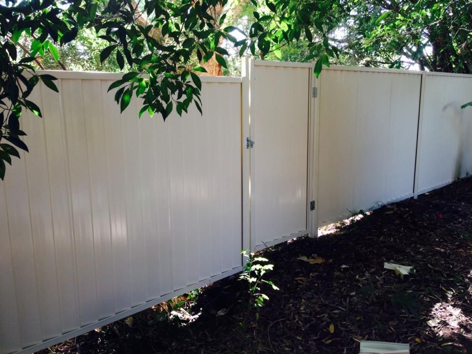 White fence along a shaded backyard with trees overhead and dark soil along the base