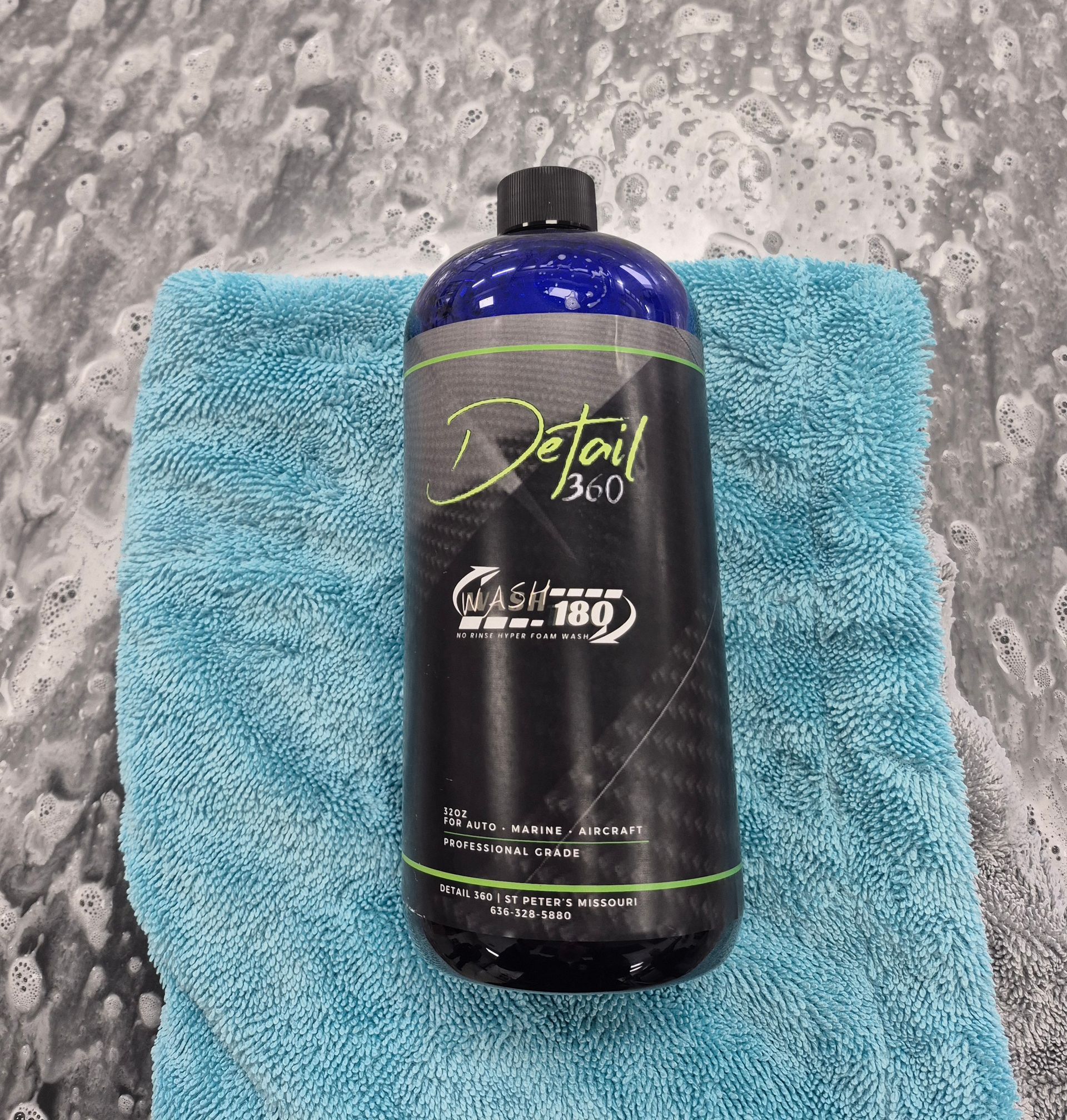 Black bottle of car wash detergent on a blue microfiber towel with soapy water in the background.