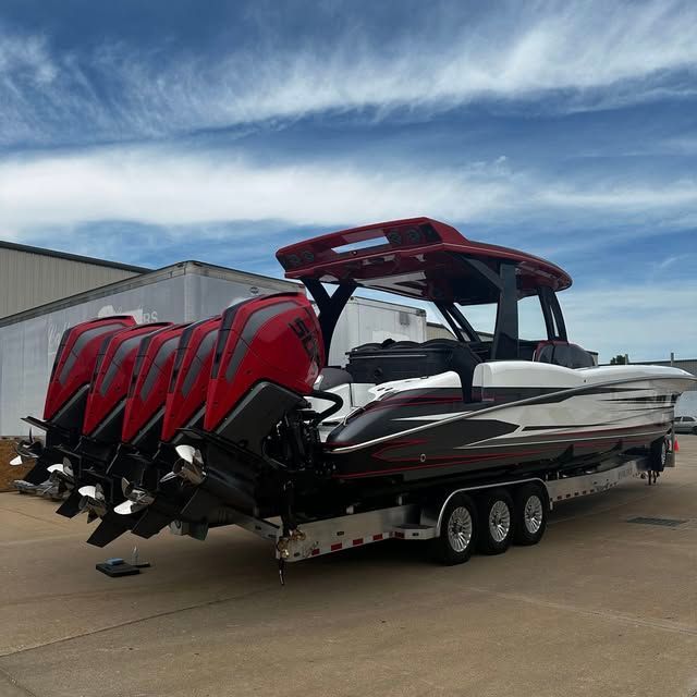 Boat with five red outboard engines on a trailer, parked on a concrete surface against a blue sky.