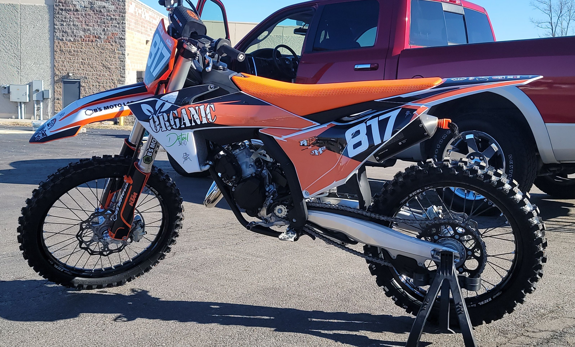 Orange and black dirt bike with the number 817 parked near a red pickup truck.