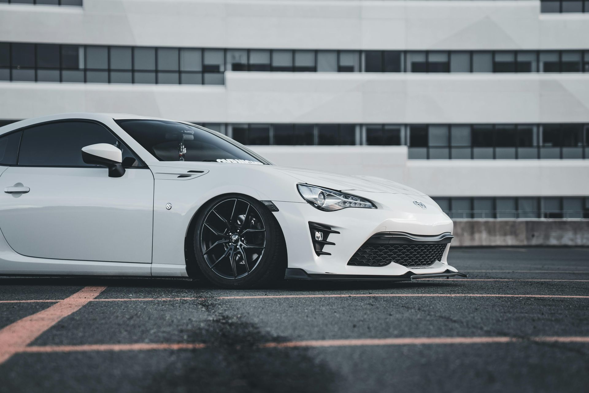 White sports car parked in front of a modern building with black rims and tinted windows.