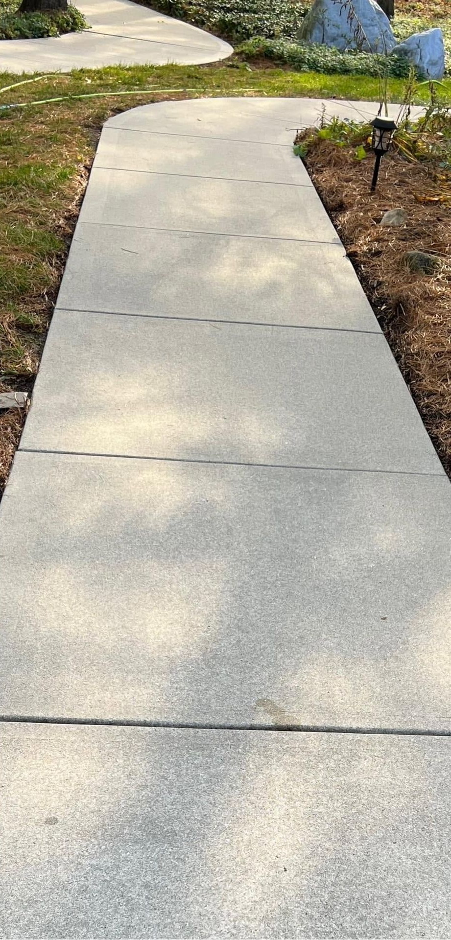 Concrete sidewalk curving through a landscaped yard with grass, shrubs, and a pond nearby