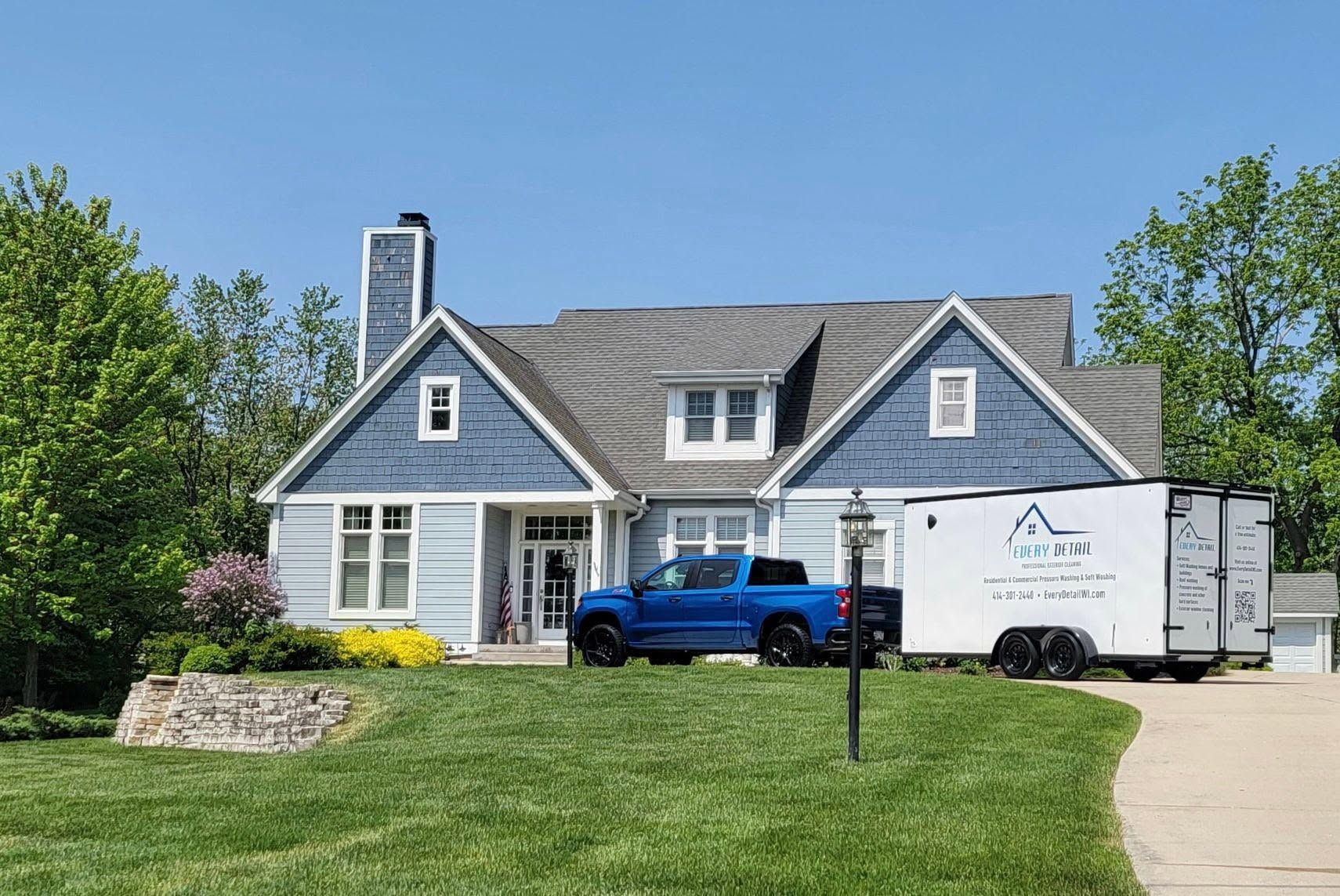 Blue house with white trim, blue pickup truck, and white moving trailer in a sunny suburban driveway.