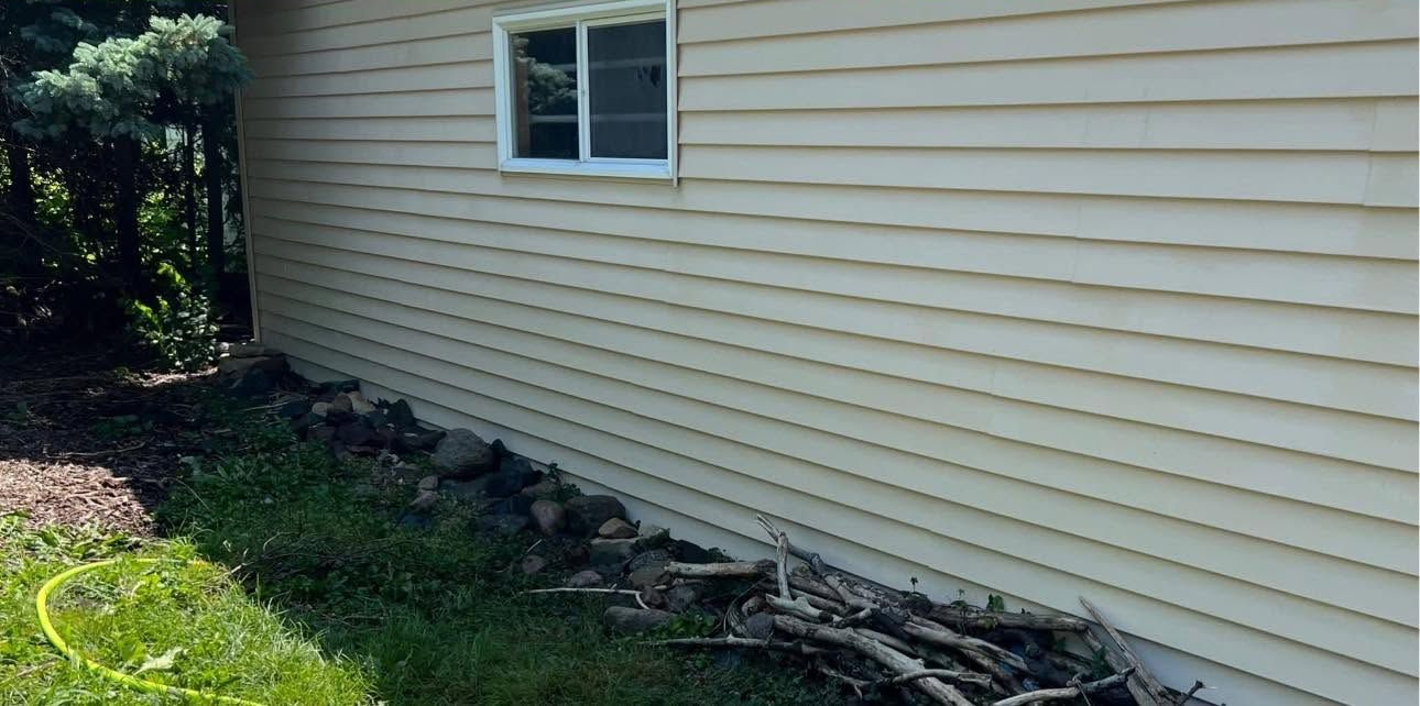 Side of a beige house with a small window and damaged siding near the foundation beside a grassy yard.