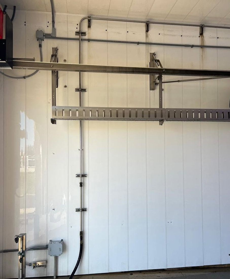 White industrial wall with a long horizontal metal rail, cables, and a mounted electrical box in a workshop-like space