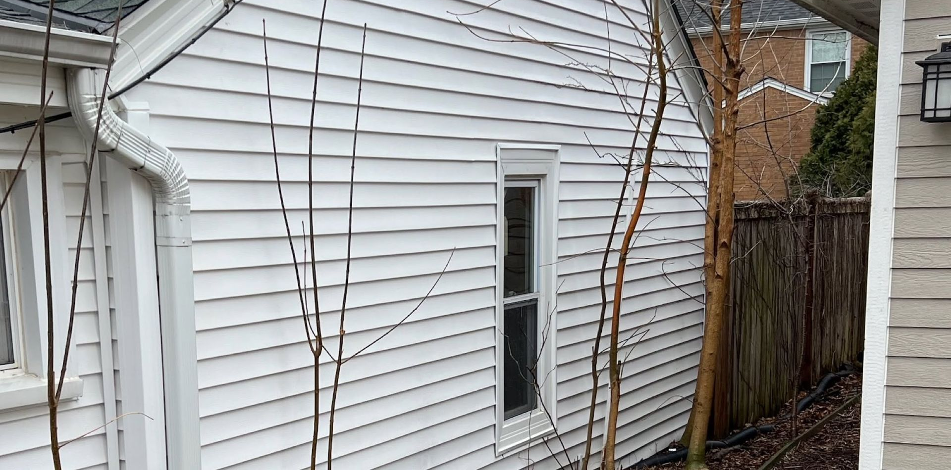 Side view of a white house with vinyl siding, a window, outdoor cables, and a narrow walkway beside a fence