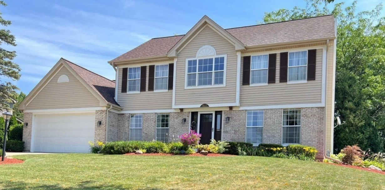 Suburban two-story house with beige siding, brick accents, two-car garage, and a green lawn