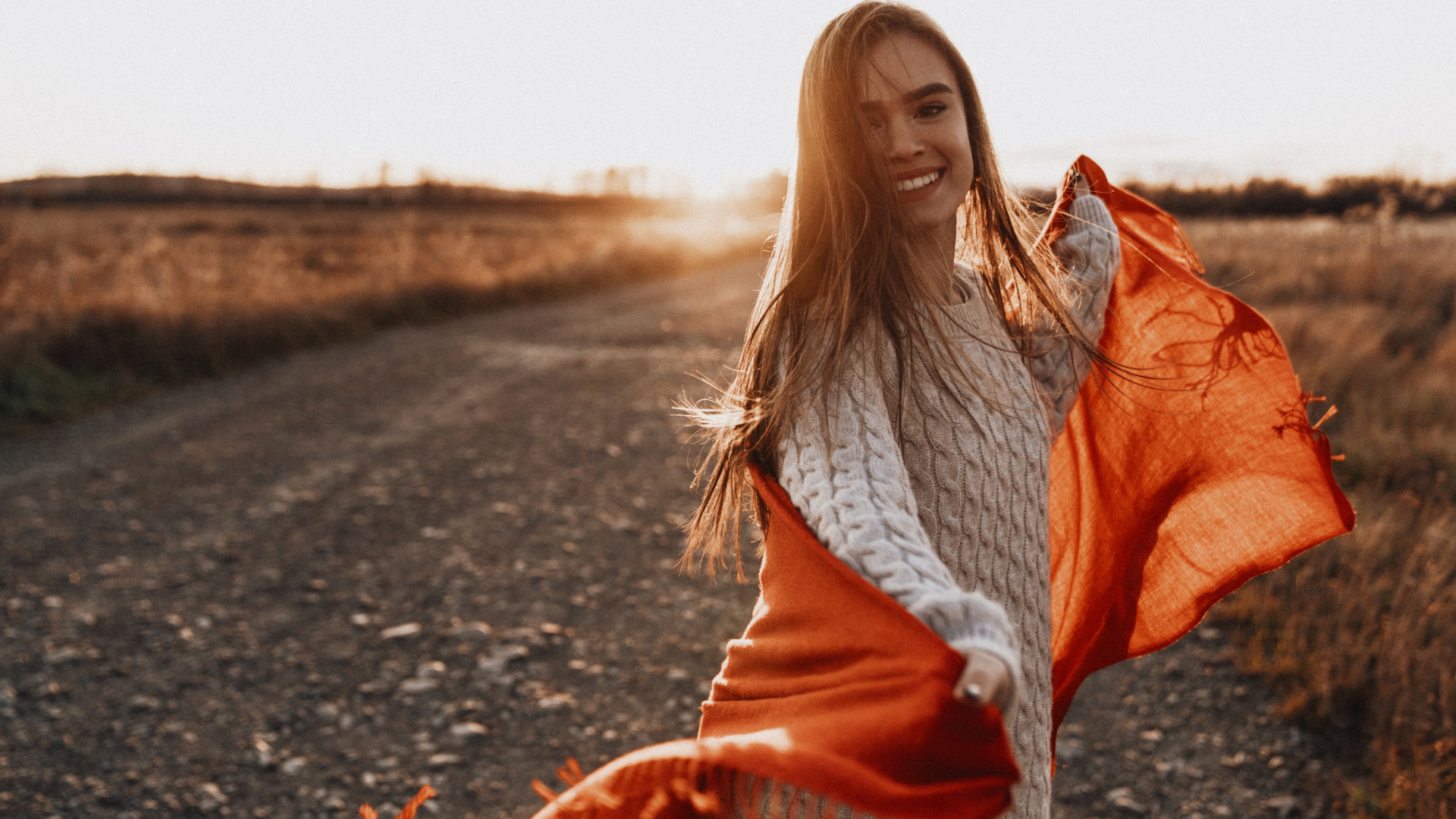 A woman is standing on a dirt road holding an orange scarf around her neck.