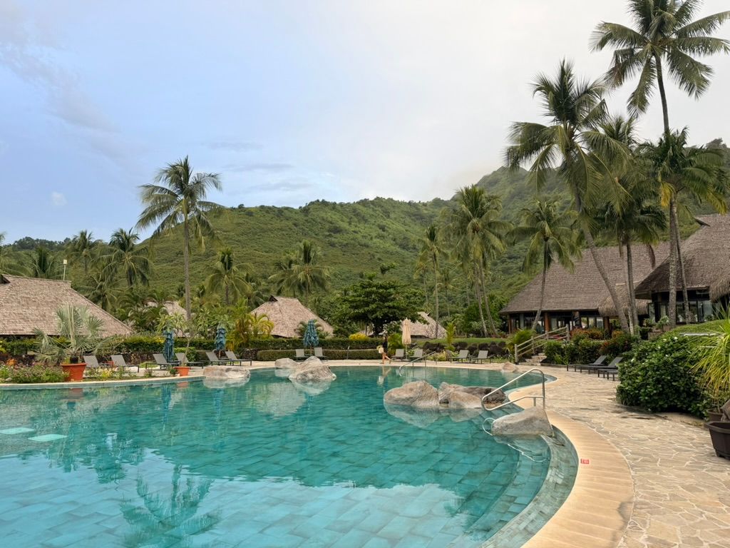 Tropical resort pool with clear blue water, palm trees, thatched huts, and lush green hills in the background