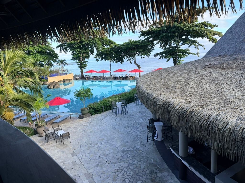 Tropical resort pool with red umbrellas, palm trees, and thatched-roof huts overlooking the water
