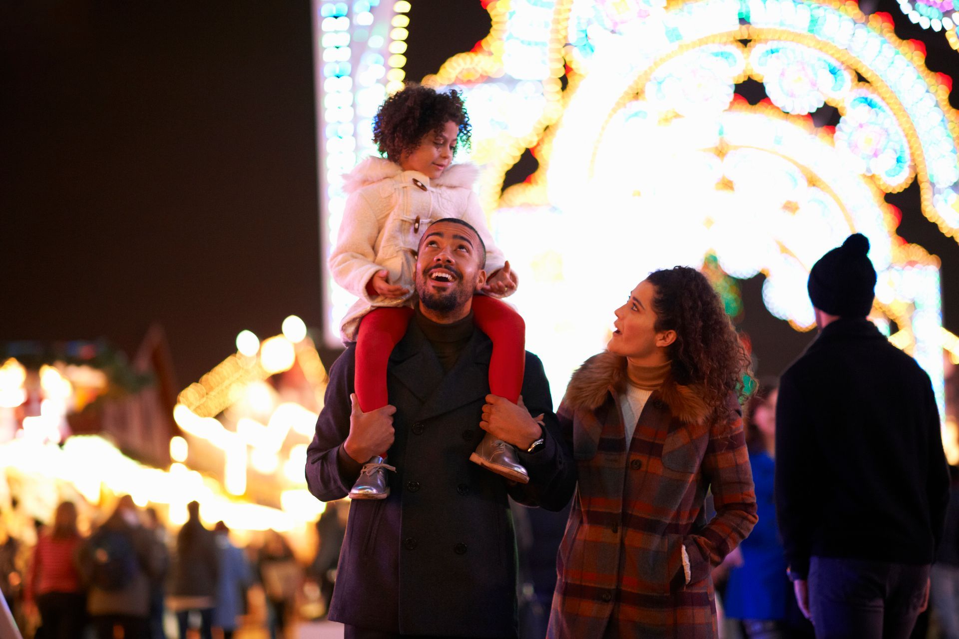 A man is carrying a little girl on his shoulders at a carnival.
