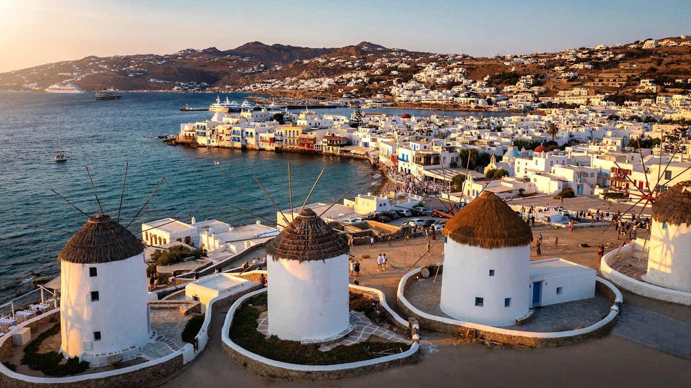 Whitewashed windmills overlook the blue sea and a town with white buildings under a sunset in Mykonos, Greece.