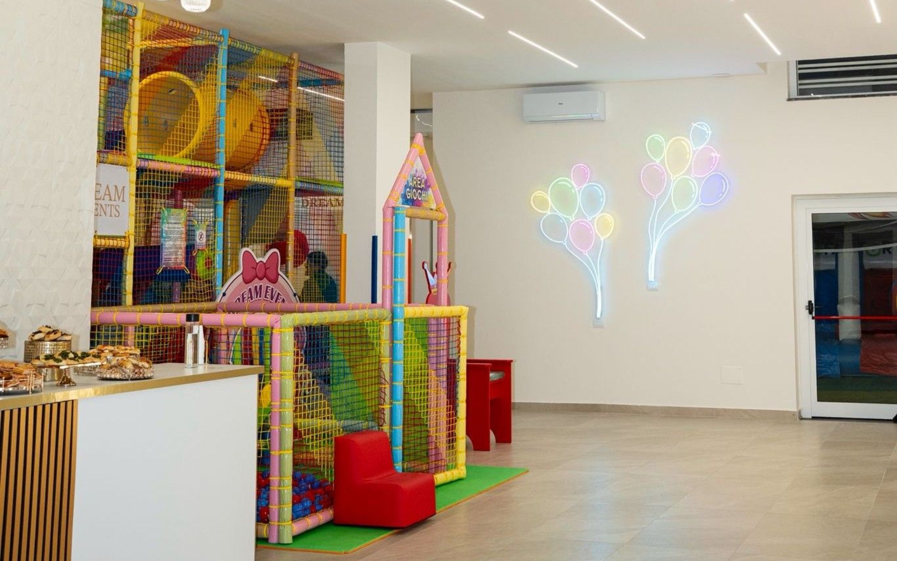 Un'area giochi con una colorata struttura per arrampicarsi, una seduta imbottita rossa e un disegno di palloncini al neon.