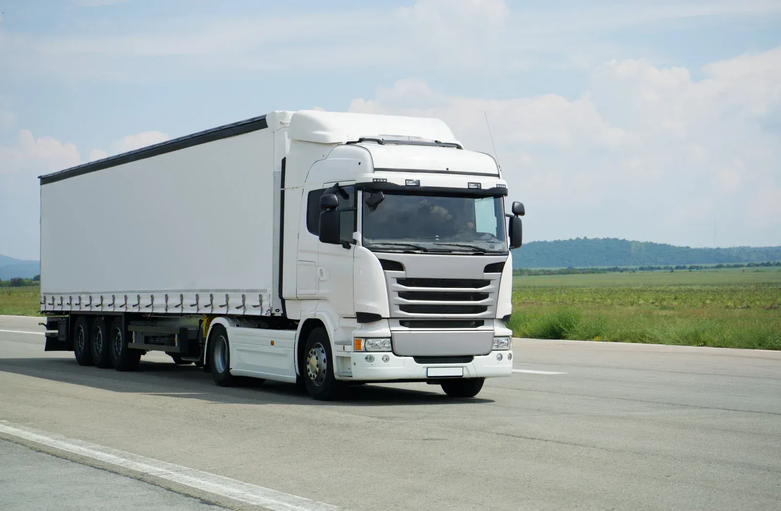 White semi-truck driving on a paved road with a blue sky and green fields in the background.