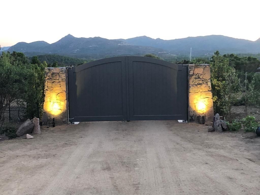 Un grande cancello nero con pilastri in pietra illuminati si erge su un vialetto di accesso a una zona rurale, con le montagne sullo sfondo.