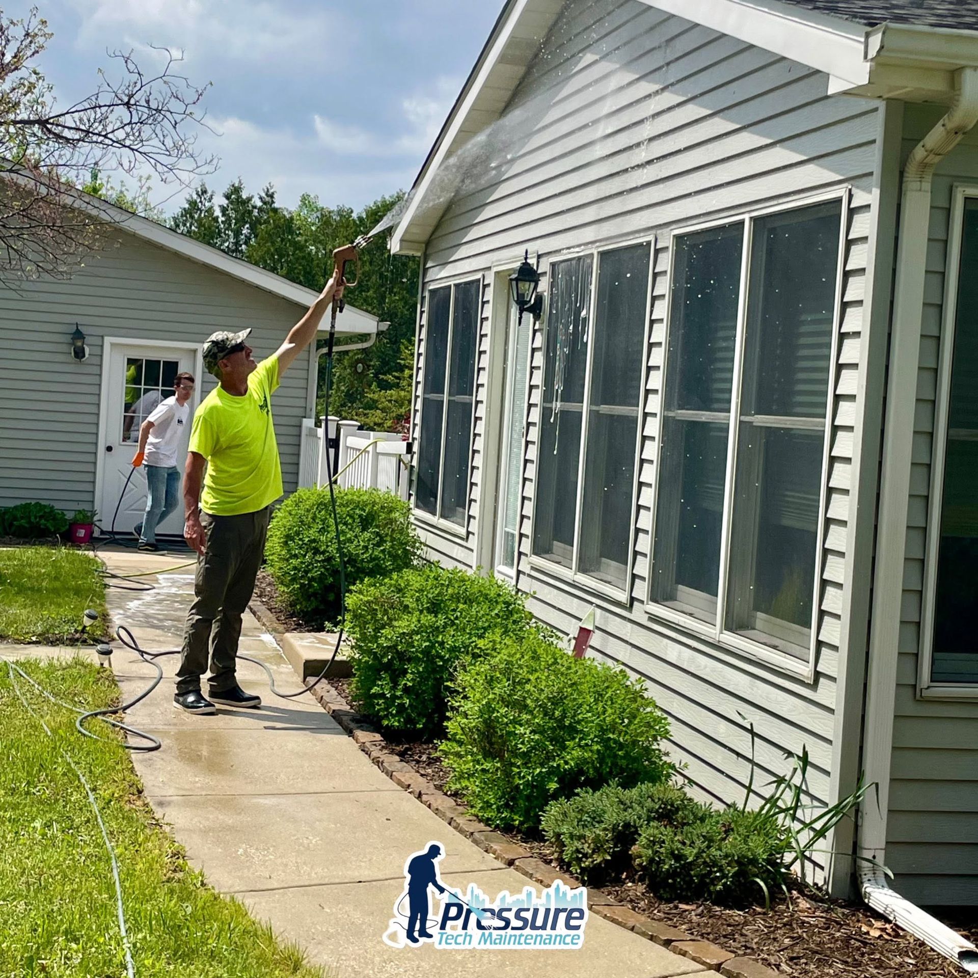 Worker pressure-washing a house exterior beside trimmed bushes on a sunny day