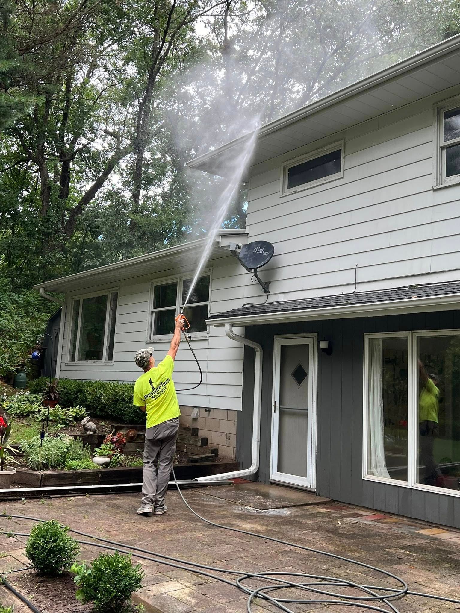 Worker pressure-washes the side of a house, spraying water up toward the roofline.