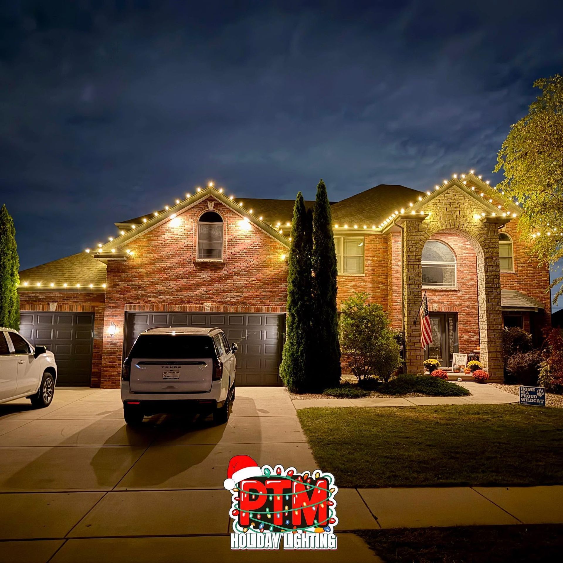 Brick house at night outlined with warm white Christmas lights, with two cars parked in the driveway.
