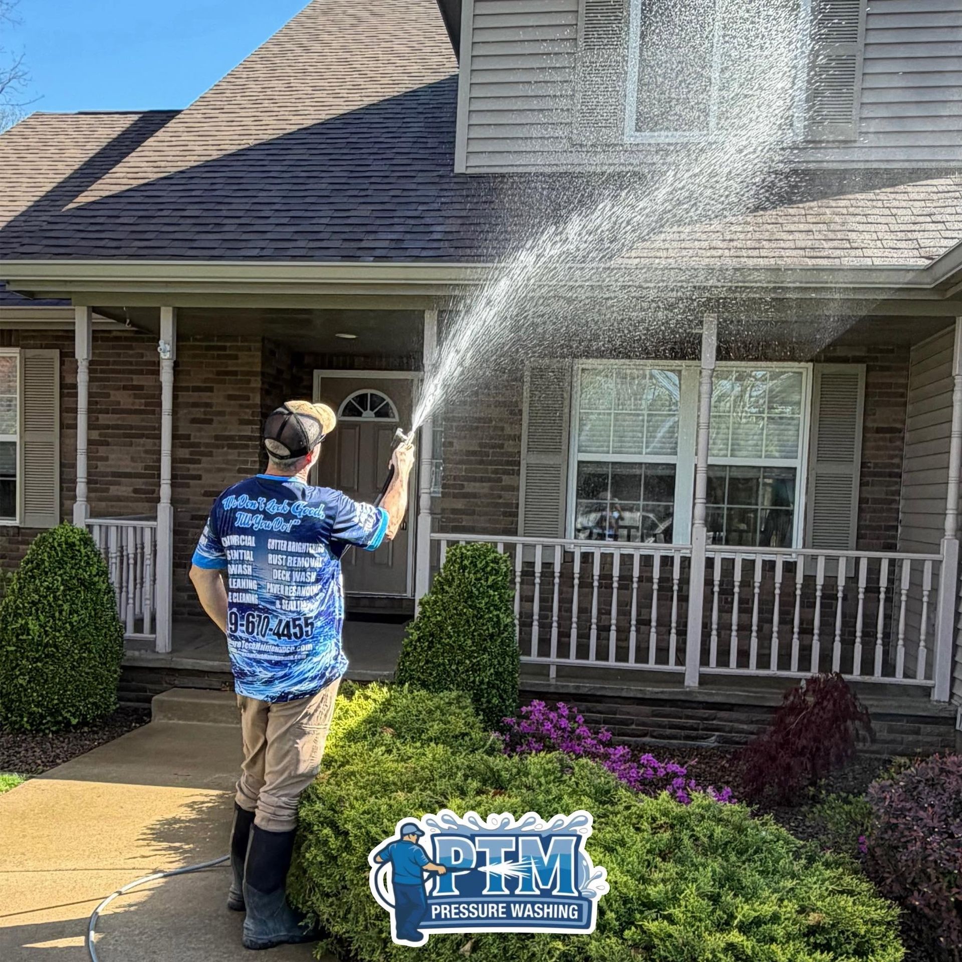 Worker spraying water on a brick house roof and siding, with a bright blue sky and front porch.