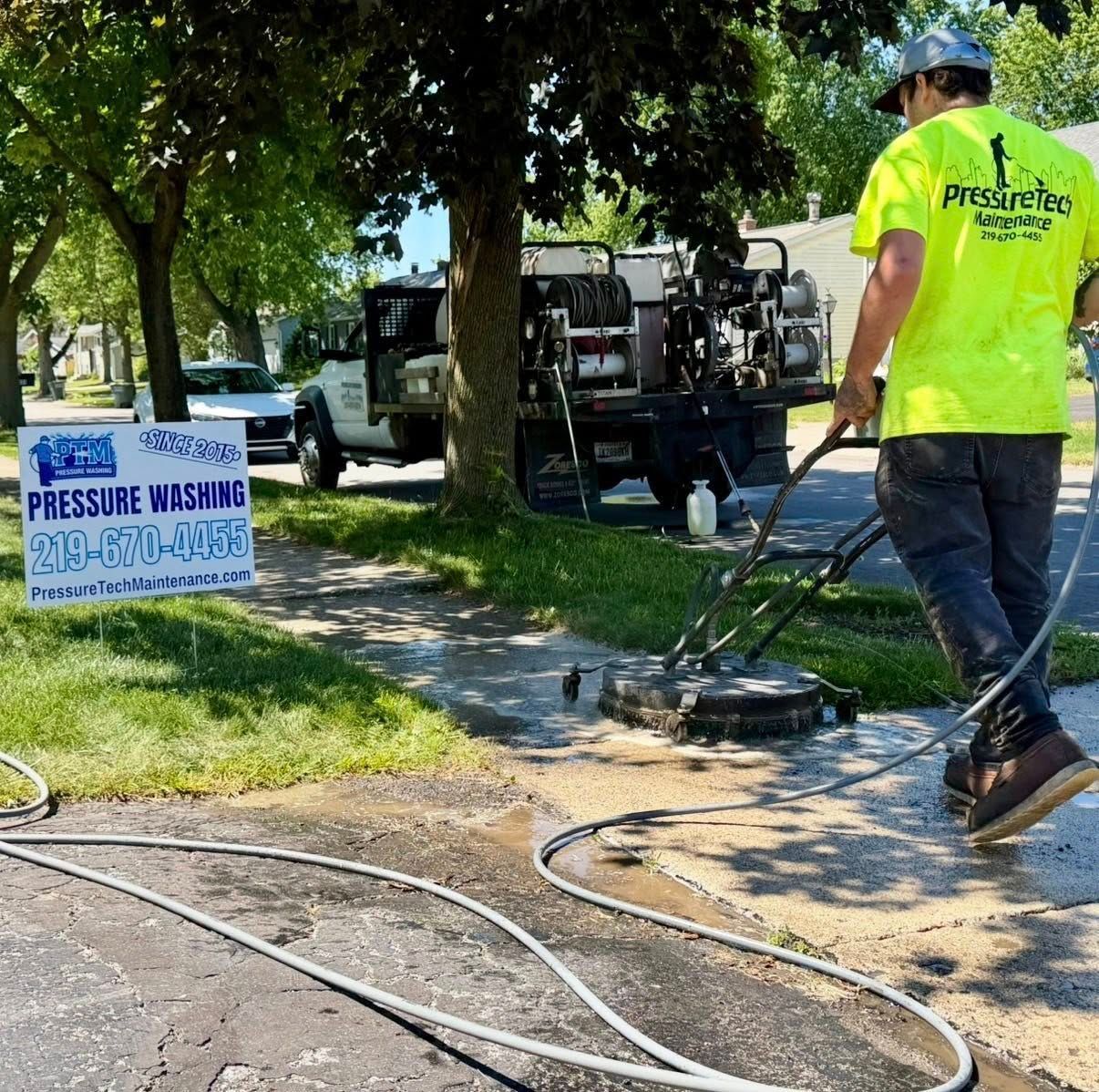 Worker pressure-washing a sidewalk beside a truck and “Pressure Washing” sign on a sunny street