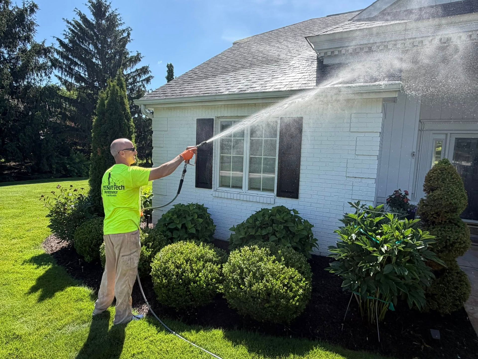 Worker pressure-washing a white house exterior and roof on a sunny day