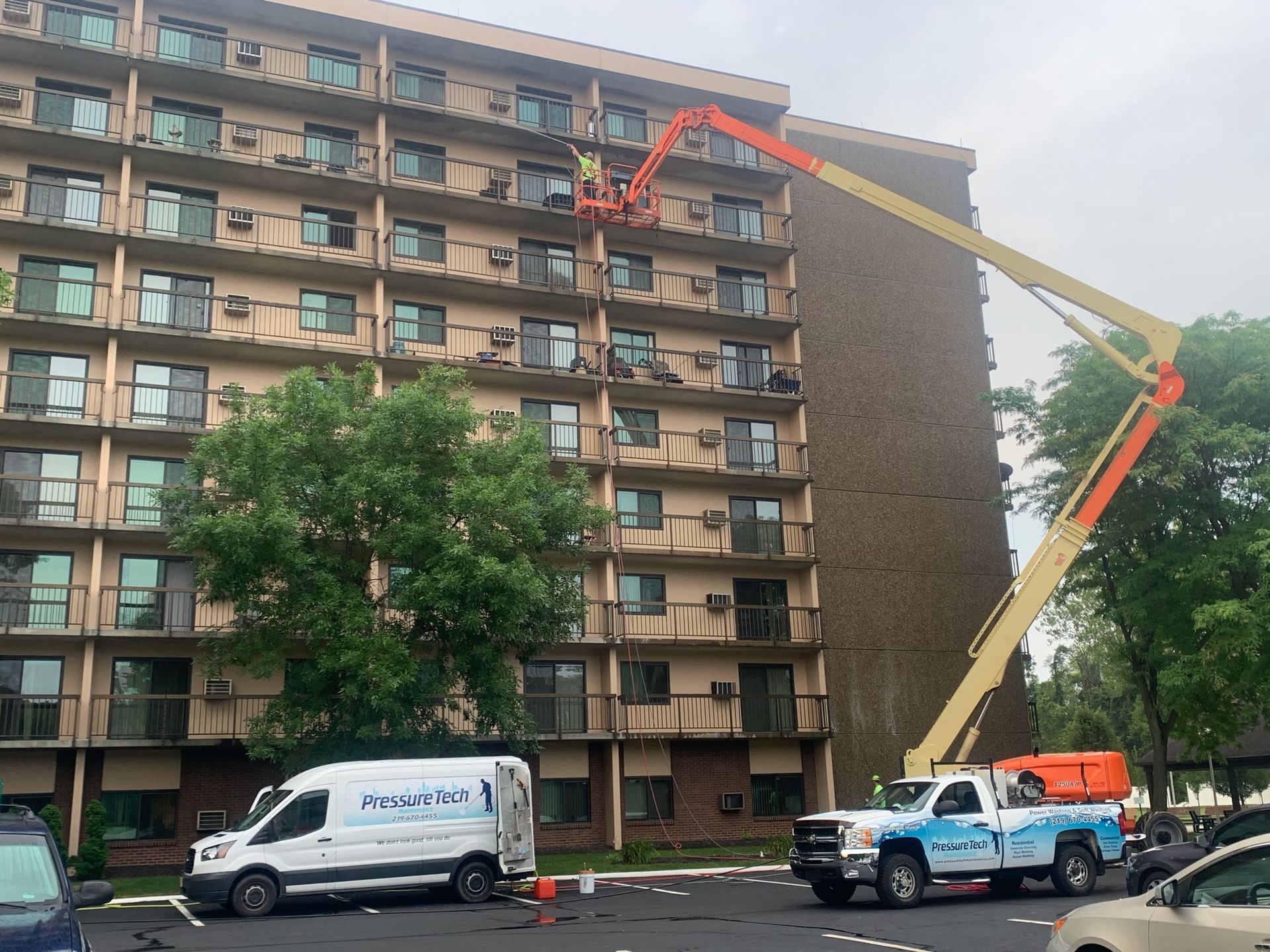 Cherry picker truck lifting a worker to the upper floors of a brown apartment building beside a tree.