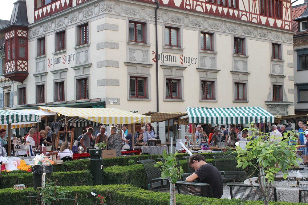 Ein Mann sitzt an einem Tisch vor einem Gebäude, davor befindet sich ein Markt.