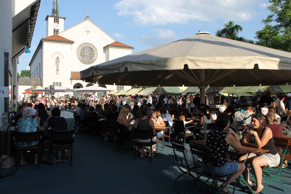 Vor einer Kirche sitzt eine Menschenmenge an Tischen unter Sonnenschirmen.
