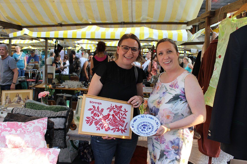 Zwei Frauen stehen nebeneinander auf einem Markt und halten ein Bild und einen Teller in den Händen.