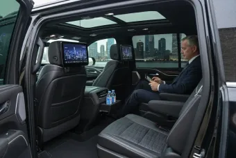 Man sits in the back of a black SUV, using a tablet. Two screens are on the headrests. City skyline visible outside.