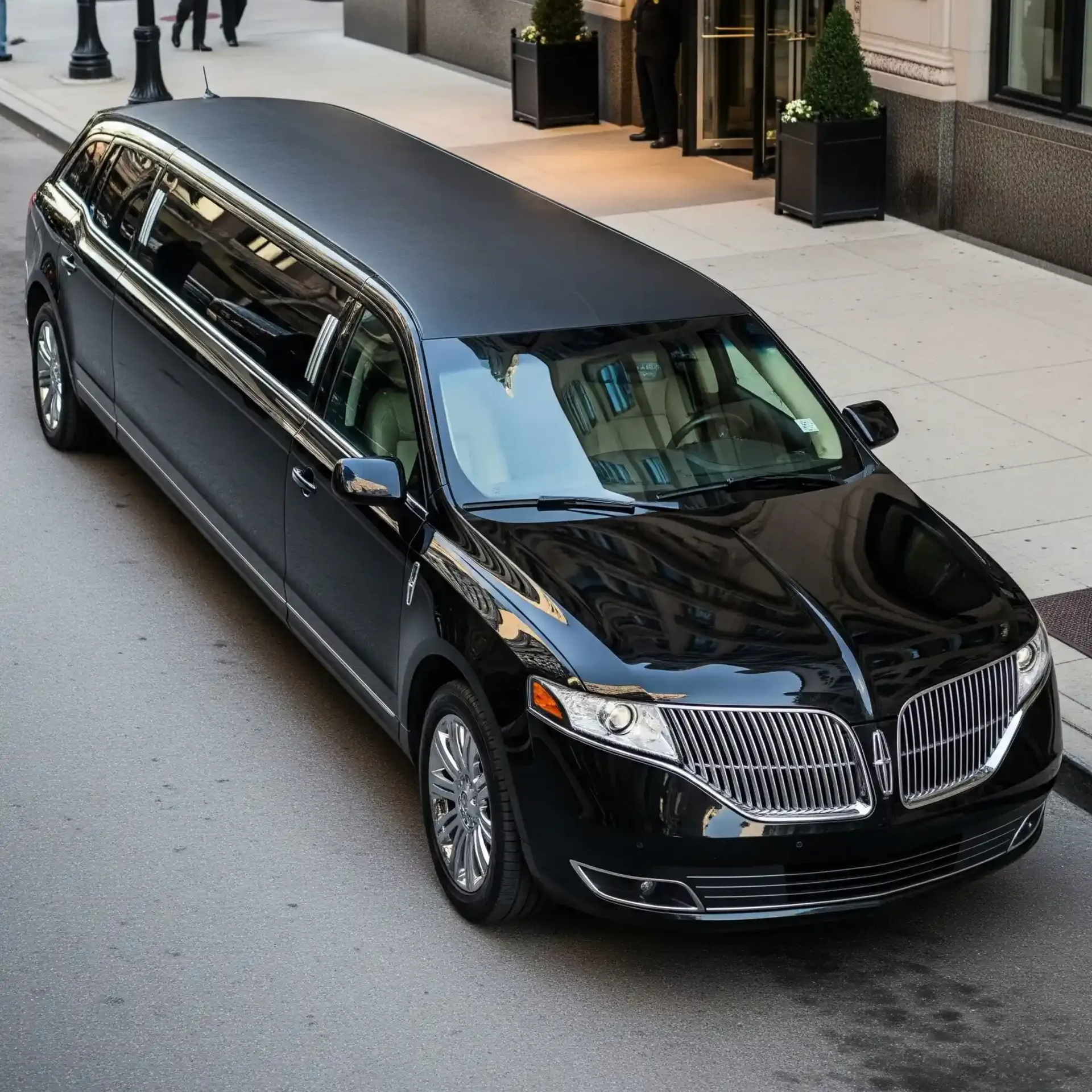 Black Lincoln limousine parked in front of a building with a revolving door. Two people in suits and a doorman are present.