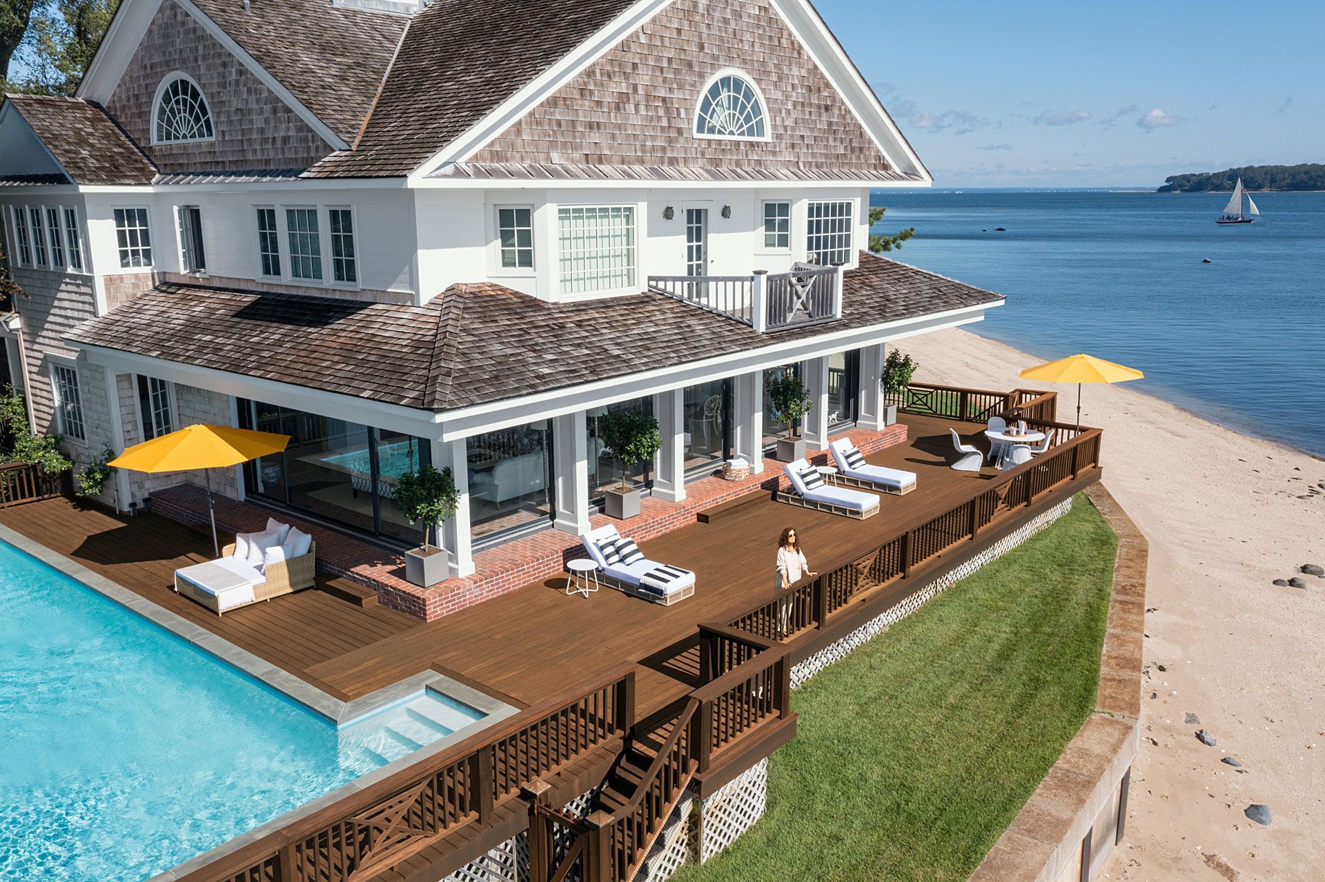 A white, multi-story coastal house with a pool deck, patio furniture, and a grassy lawn overlooking the ocean.