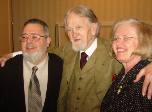 Three people pose together indoors: two men and a woman. They smile, with one man's arm around the other's shoulder.