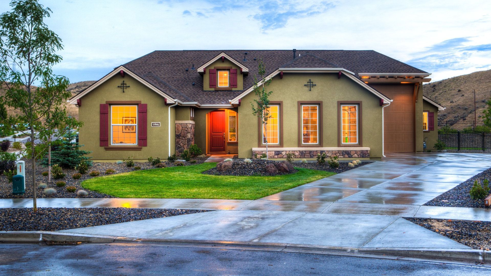 Olive-green house with red door and shutters, wet driveway, green lawn. Cloudy sky, mountains in the background.