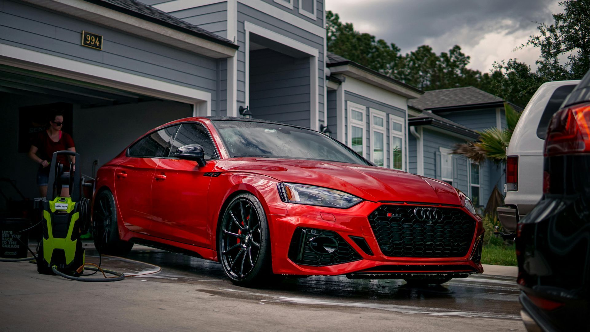 Red Audi RS5 being washed in front of a blue house with a person and pressure washer visible.