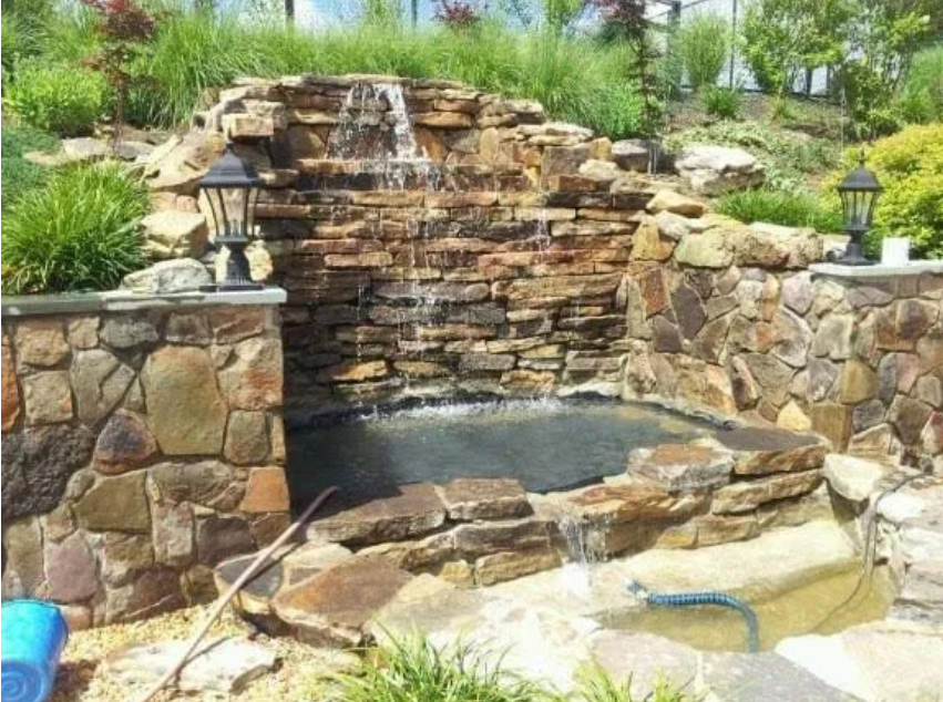 Stone backyard waterfall cascading into a small pond with surrounding rocks and greenery