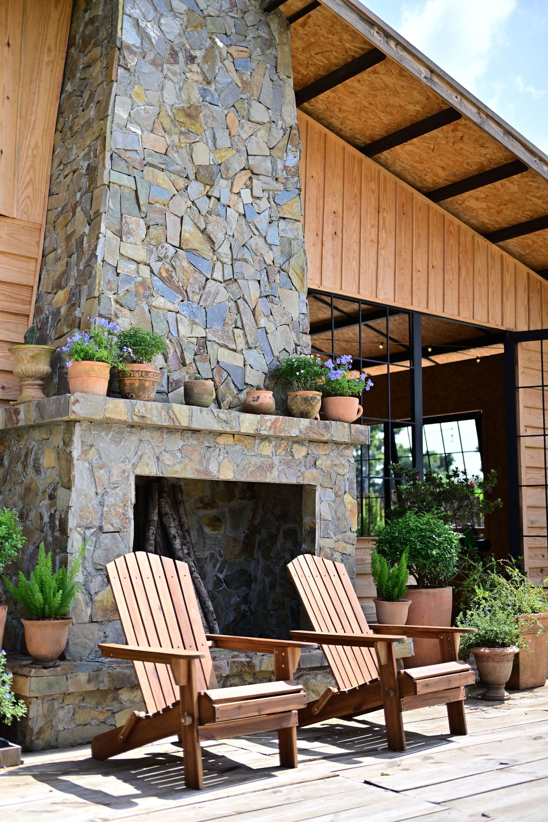 Rustic stone fireplace with two wooden chairs and potted plants on a sunny patio