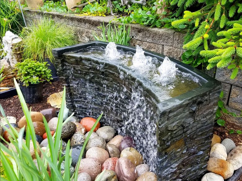 Small garden fountain with cascading water over stone, surrounded by pebbles and greenery