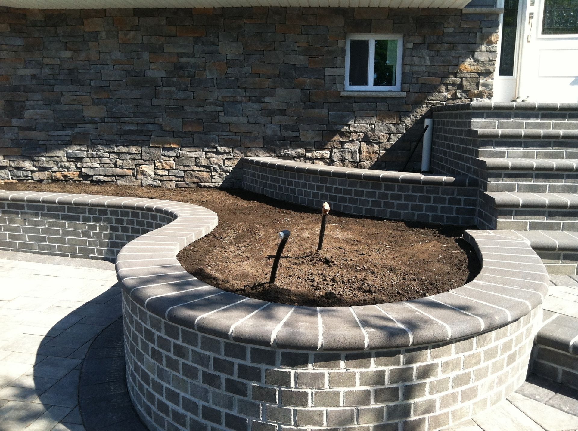 Circular brick planter with bare soil and a small pipe, set beside stone steps and a brick wall outdoors