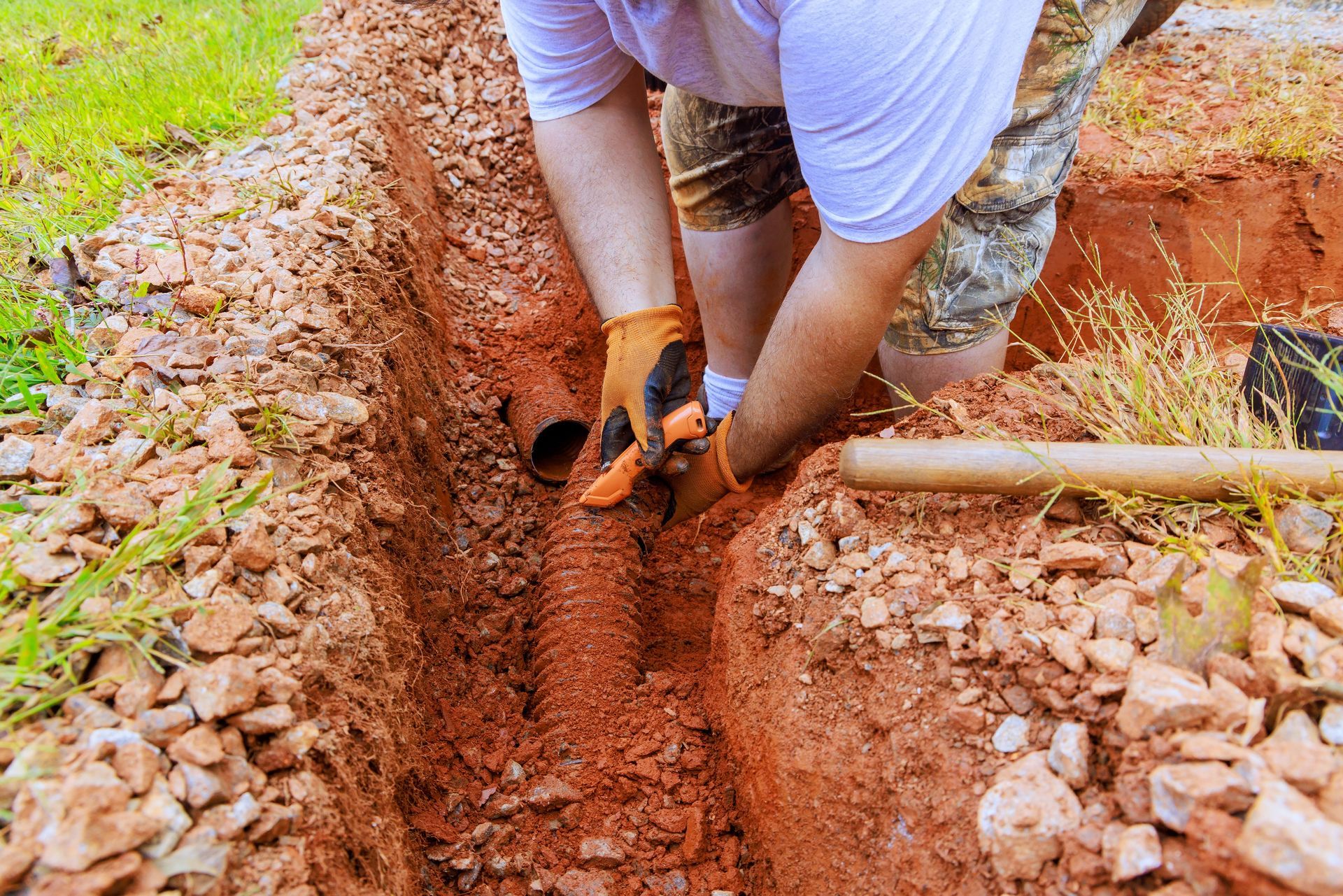 Person digging a trench in red clay with a shovel and blue gloves outdoors