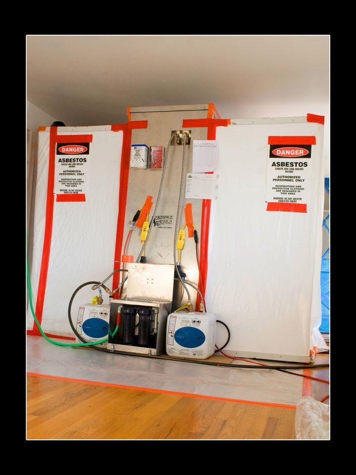 Containment setup for asbestos removal: white panels, orange tape, warning signs, equipment, hardwood floor.