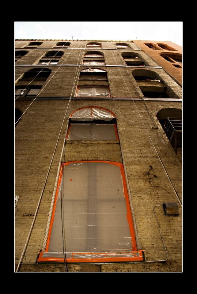 Brick building with arched windows, some boarded up. Orange frames and cables. Low angle view.