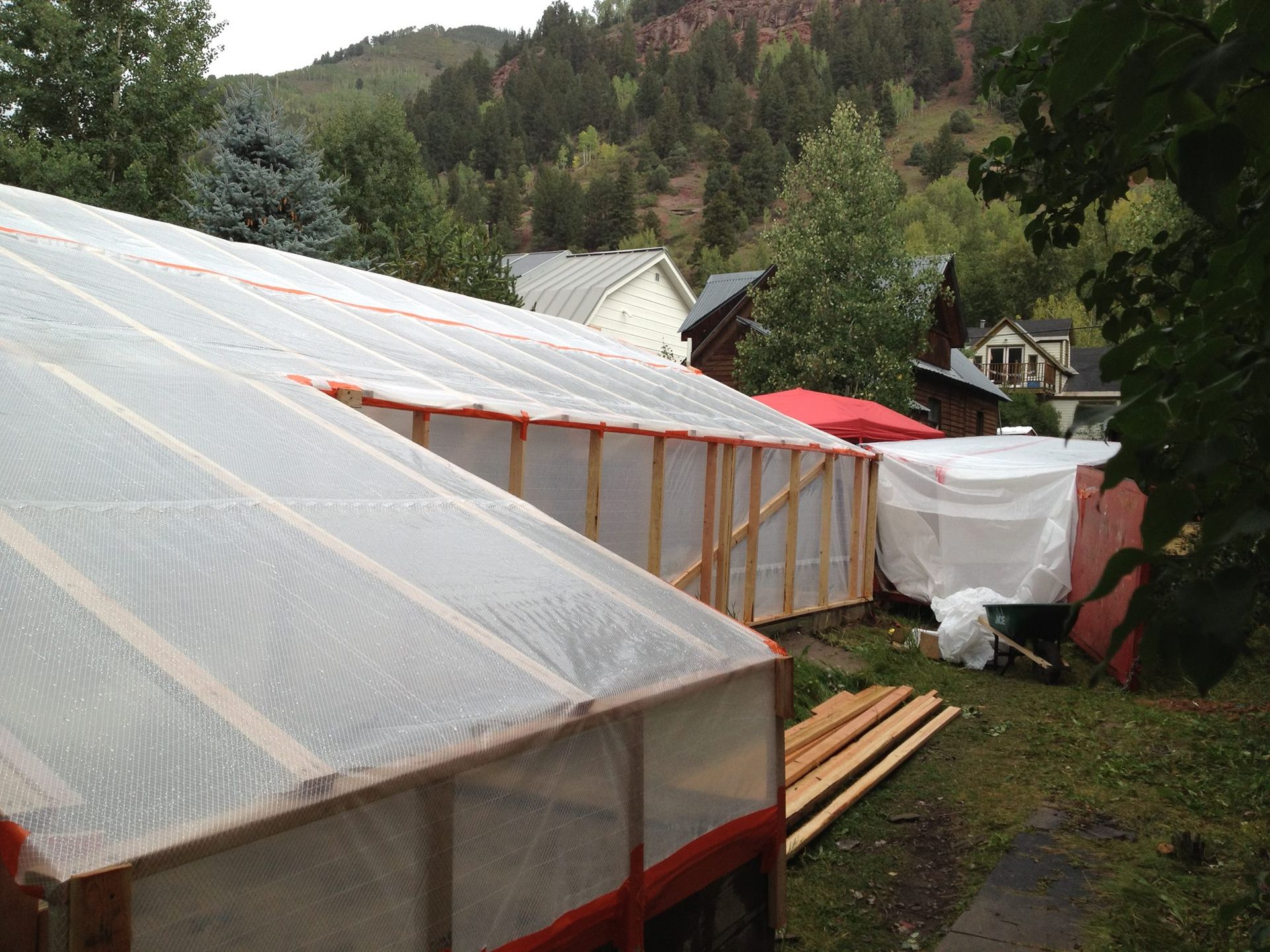 Wooden-framed structures covered in plastic sheeting, potentially greenhouses, in a yard with a backdrop of a mountain and buildings.