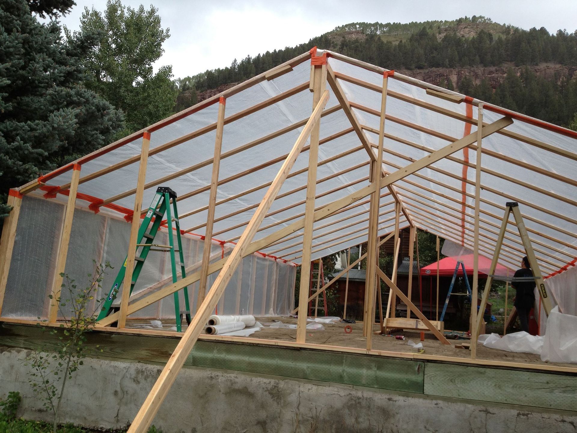 A partially constructed greenhouse with wooden frame and plastic sheeting, in a natural setting.