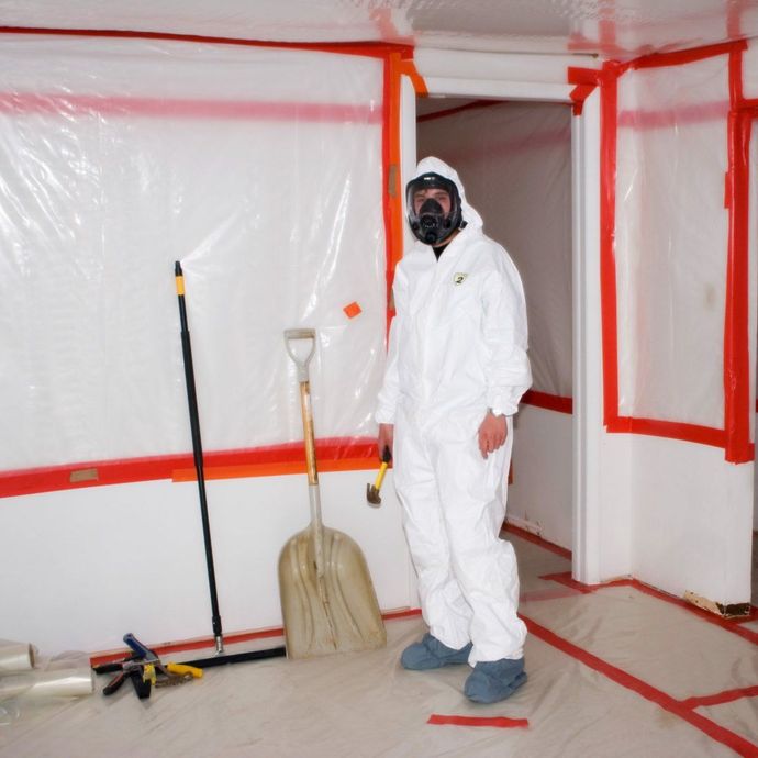 Person in protective suit and respirator stands in a room sealed with plastic sheeting and red tape; tools lie nearby.