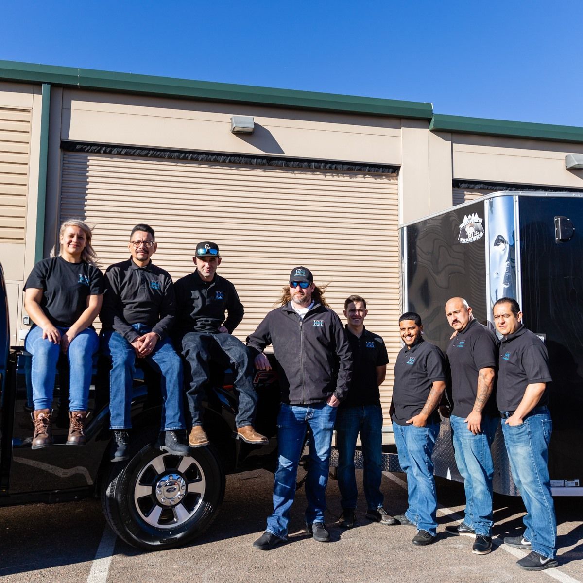 Group of people posing outdoors in front of a building and vehicles, wearing casual clothing.
