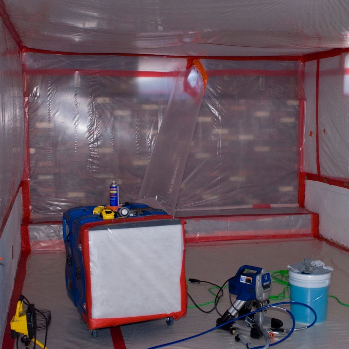 Interior of a plastic-lined room set up for painting. Equipment, tools, and a blue container are inside.