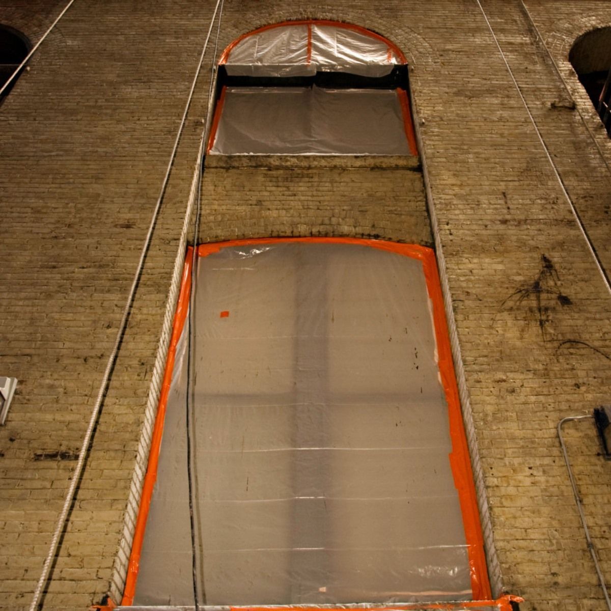 Tall, arched windows in brick building, covered in plastic sheeting with orange tape.