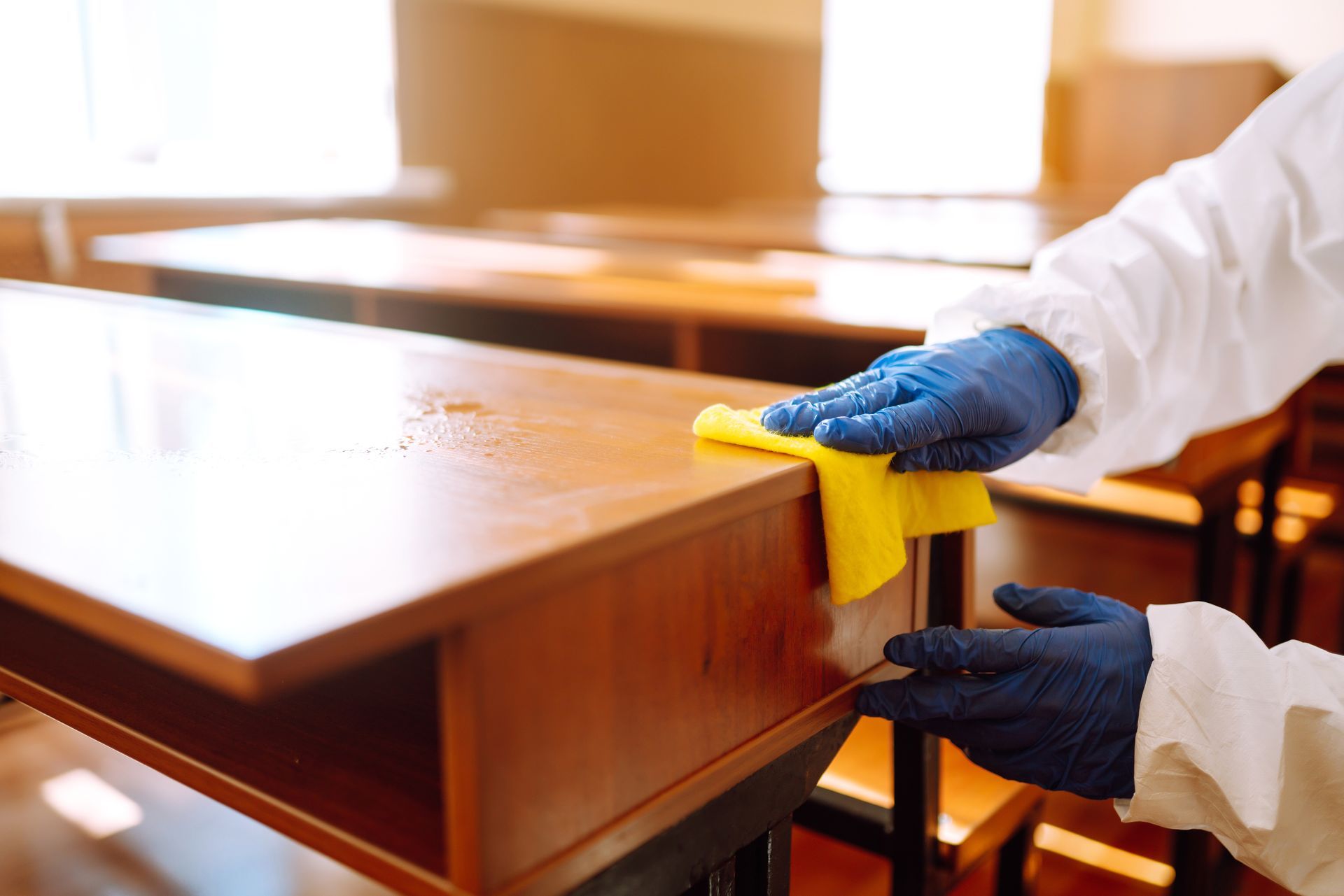 Person in protective suit and gloves wiping a wooden desk with a yellow cloth.